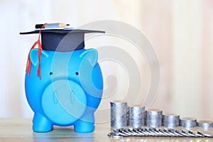 Graduation hat on blue piggy bank with stack of coins money on wooden background, Saving money for education concept