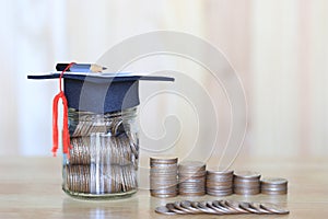 Graduation hat on the glass bottle with Stack of coins money on wooden background, Saving money for education concept