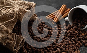 Grains of coffee pour out from a white coffee cup on a dark background from a bag with cinnamon sticks