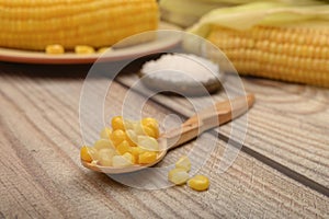 Grains of sweet corn on a wooden spoon on a wooden table, in the background boiled corn, coarse salt and untreated ears of corn.