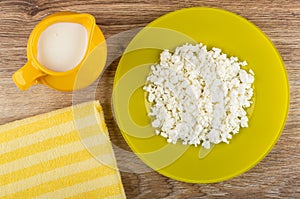 Grainy defatted cottage cheese in yellow plate, jug with yogurt, napkin on table. Top view