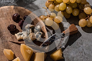 Grana padano and dorblu with olives and knife on round wooden board next to grapes on grey background.