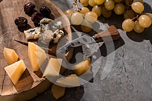 Grana padano and dorblu with olives and knife on wooden board next to grapes on grey background.