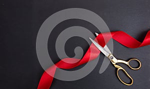 Grand opening. Top view of gold scissors cutting red silk ribbon against black background