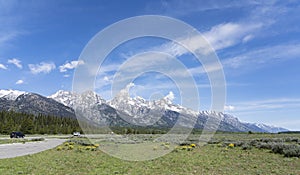 Grand Teton National Park is viewed from a green grass meadow. Mountains and clouds touch. Cars drive. Sky is blue.