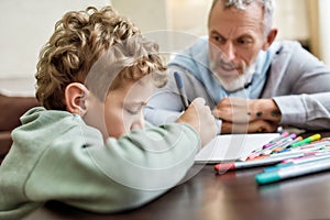 Grandad with serious face helping his little grandson drawing with colored pencils, spending time at home