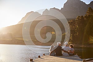 Grandchildren With Grandparents Sitting On Wooden Jetty By Lake