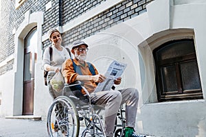 Granddaughter pushing senior man in wheelchair on street. Buying newspaper in newsstand. Female caregiver and elderly