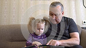 Granddaughter is reading a book with grandfather. The girl frowns at the book and listens carefully to Grandfather