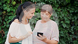 Granddaughter shows old grandmother something in smartphone, teaches her to handle with modern gadget and technology