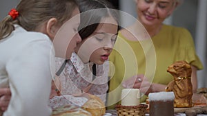 Granddaughters blowing out candle on table with grandmother smiling sitting at background. Positive girls and happy