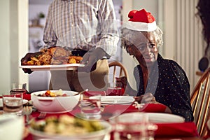 Grandfather Carrying Turkey As Multi-Generation Family Sit Down To Eat Christmas Meal Together