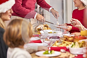 Grandfather Carving Turkey As Multi-Generation Family Sit Down To Eat Christmas Meal Together