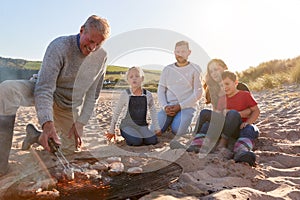 Grandfather Cooking As Multi-Generation Family Having Evening Barbecue Around Fire On Beach Vacation