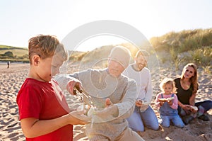 Grandfather Cooking As Multi-Generation Family Having Evening Barbecue Around Fire On Beach Vacation