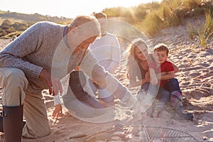 Grandfather Cooking As Multi-Generation Family Having Evening Barbecue Around Fire On Beach Vacation