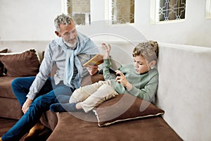 Grandfather giving book to his little grandson while child playing video games on smartphone, they sitting on sofa at