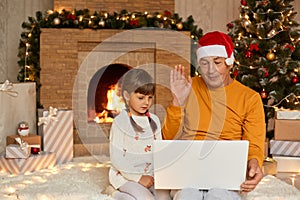 Grandfather and granddaughter having video on notebook, waving hands to camera, congratulating somebody with new year eves,