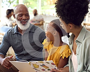 Grandfather With Granddaughter And Mother Reading Book At Home With Multi-Generation Family Behind