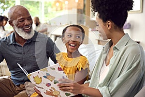 Grandfather With Granddaughter And Mother Reading Book At Home With Multi-Generation Family Behind