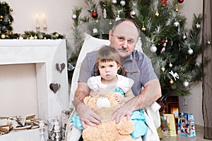 Grandfather and granddaughter portrait in Christmas interior. Little girl sitting behind older father with Teddy bear