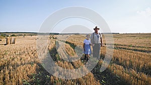 Grandfather and granddaughter walking across the field with haystacks. Farmer grandfather teaches the younger generation