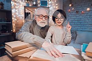 Grandfather and grandson are doing homework at night at home. Granddad is helping boy.