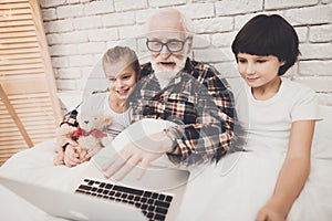 Grandfather, grandson and granddaughter at home. Grandpa and children are lying using laptop.