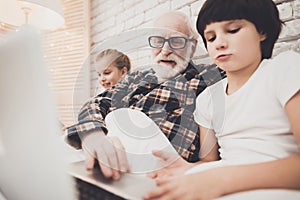 Grandfather, grandson and granddaughter at home. Grandpa and children are reading book and using laptop.