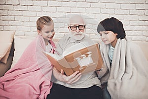 Grandfather, grandson and granddaughter at home. Grandpa and children are watching photos in album.