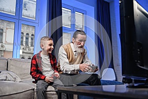 Grandfather and grandson are playing exciting video games on computer with gamepads at home.