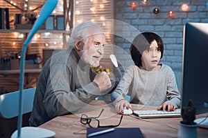 Grandfather and grandson are playing games on computer at night at home. Granddad is cheering for boy.