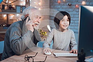 Grandfather and grandson are playing games on computer at night at home. Granddad is on phone.