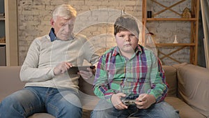 Grandfather and grandson are sitting on the couch. An old man uses a tablet, a young fat guy plays on the console game