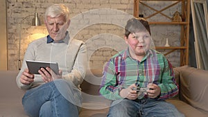 Grandfather and grandson are sitting on the couch. An old man uses a tablet, a young fat guy plays on the console game