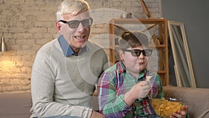 Grandfather and grandson are sitting on the couch and watching a 3D movie in 3d glasses, eating chips, laughing, smiling