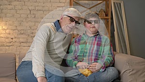 Grandfather and grandson are sitting on the couch and watching a 3D movie in 3d glasses, eating chips, moving, TV, show