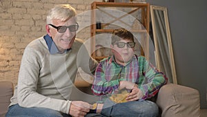 Grandfather and grandson are sitting on the couch and watching a 3D movie in 3d glasses, eating chips, smiling, TV, show
