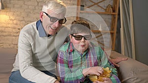 Grandfather and grandson are sitting on the couch and watching a 3D movie in 3d glasses, eating chips, speaking, dancing