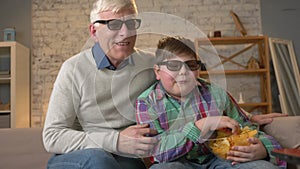 Grandfather and grandson are sitting on the couch and watching a 3D movie in 3d glasses, eating chips, TV, show. Home