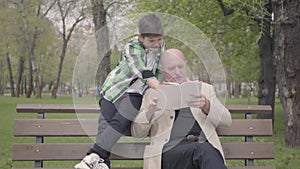 Grandfather and grandson sitting in the park on the bench, old man reading the book for the boy. The child standing with