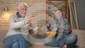 Grandfather and his grandson are sitting on the couch and watching television, eating chips, smiling. An elderly man