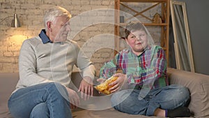 Grandfather and his grandson are sitting on the couch and watching television, fat boy offers grandfather to eat chips