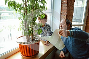 Grandfather and his grandson spending time insulated at home, having fun, caring for plants, watering