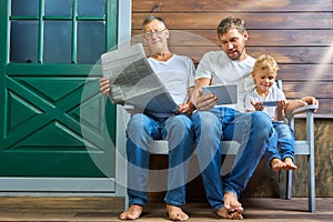 Grandfather with a newspaper, son with a tablet, grandson with a smartphone are sitting on a bench