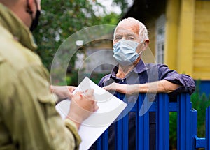 Grandfather in protective mask talking to census agent standing at fence of his country house