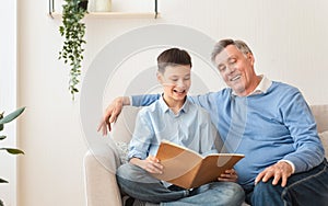 Grandfather Reading Book With Grandson Sitting On Sofa At Home