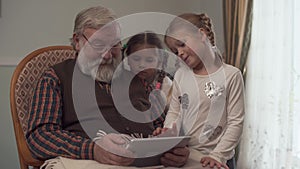 Grandfather shows his two cute granddaughters something on the tablet, swinging on a rocking chair with a plaid