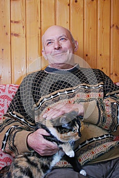 The grandfather sits on a sofa at the dacha and irons the cat sitting at it on a lap.