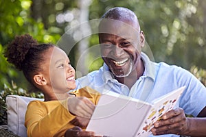 Grandfather Sitting Outdoors With Granddaughter At Home Reading Book Together